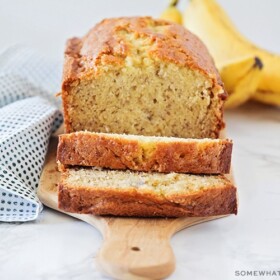 a loaf of homemade banana bread made with this easy recipe . Two slices have been cut off from one end and are laying down on the cutting board. Behind the loaf of bread is a small bunch of bananas.