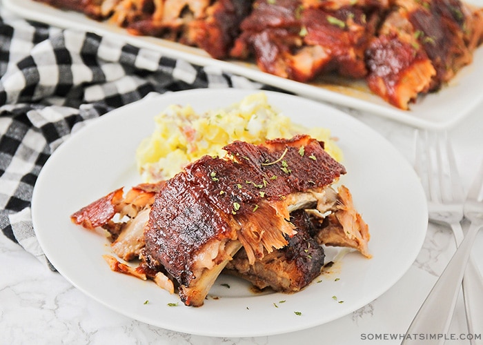 a white plate with three pork ribs and a side of potato salad. A tray of additional ribs hot off the grill are behind the plate.