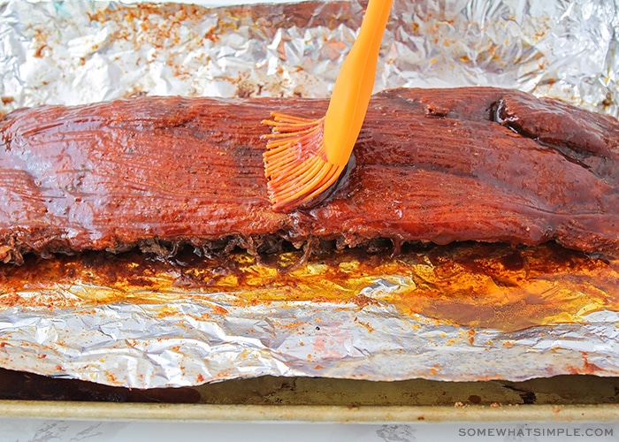 an orange basting brush spreading bbq sauce over a cooked rack of ribs