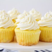 a white cake stand with several lemon cupcakes topped with a cream cheese frosting and white sprinkles. There are a couple of lemons on the counter next to the cake stand.