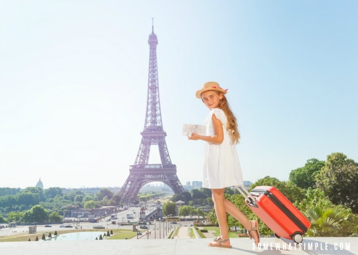 a girl in a white dress pulling a rolling suitcase in one hand and holding a packing list in the other. The Eiffel Tower is in the background.